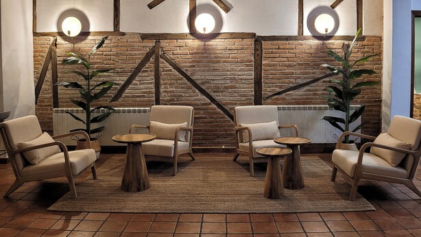 Een knus zithoekje met vier fauteuils, twee houten bijzettafeltjes en een groot geweven vloerkleed. De ruimte heeft zichtbare bakstenen muren en houten balken, met twee wandlampen.