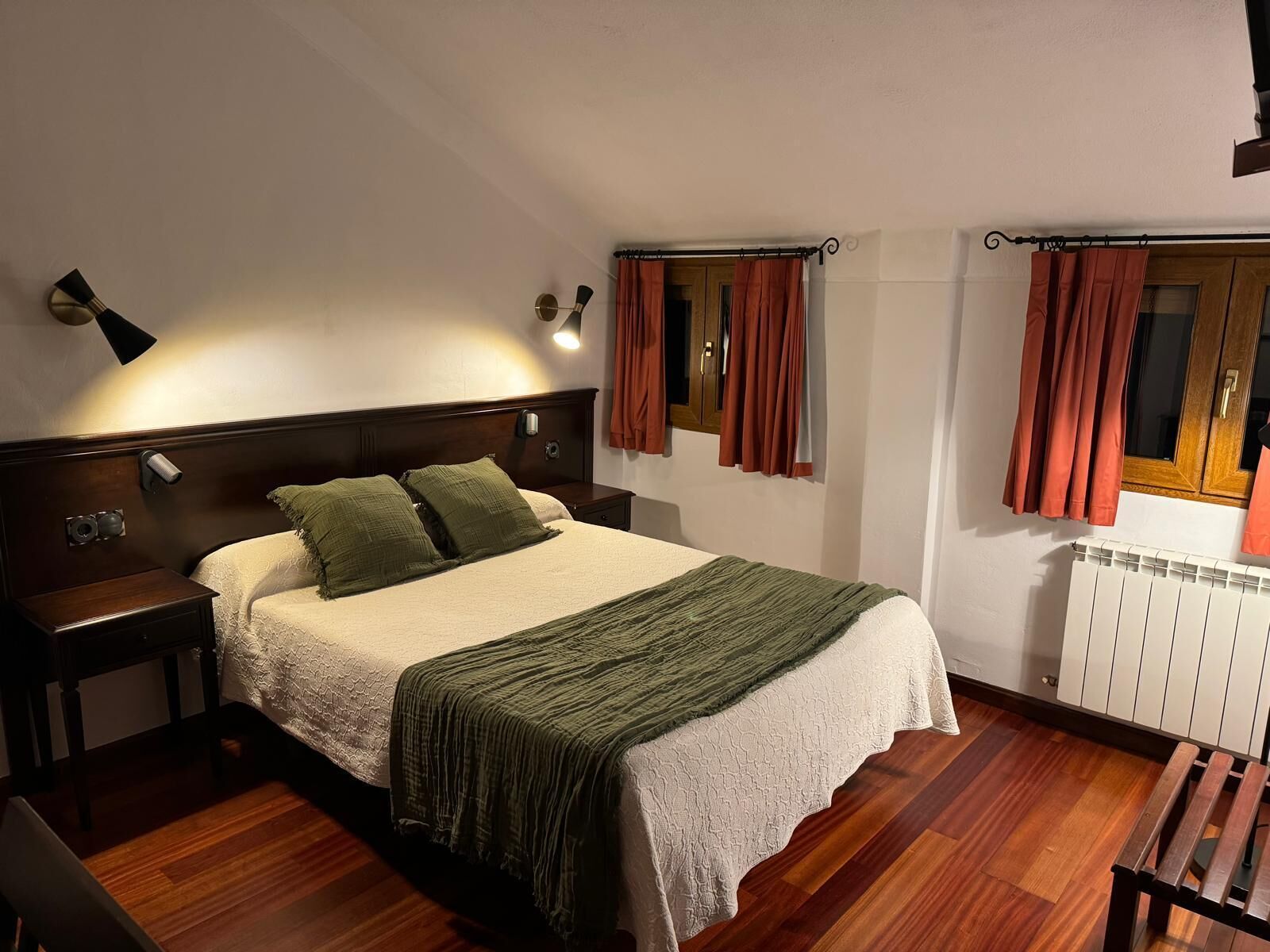A bedroom with a bed, nightstands, a wooden bench, a radiator, and curtains.