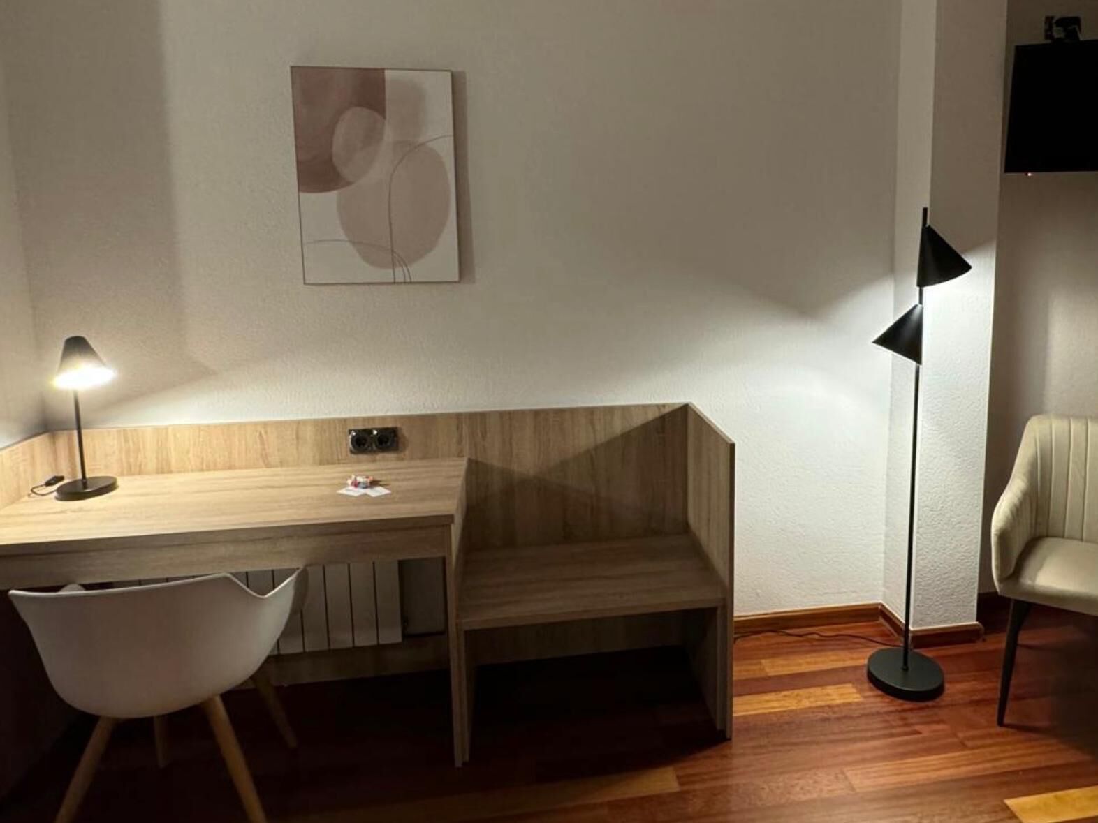 A modern, minimalist room with a wooden desk, a white chair, a floor lamp, and a wall-mounted artwork.