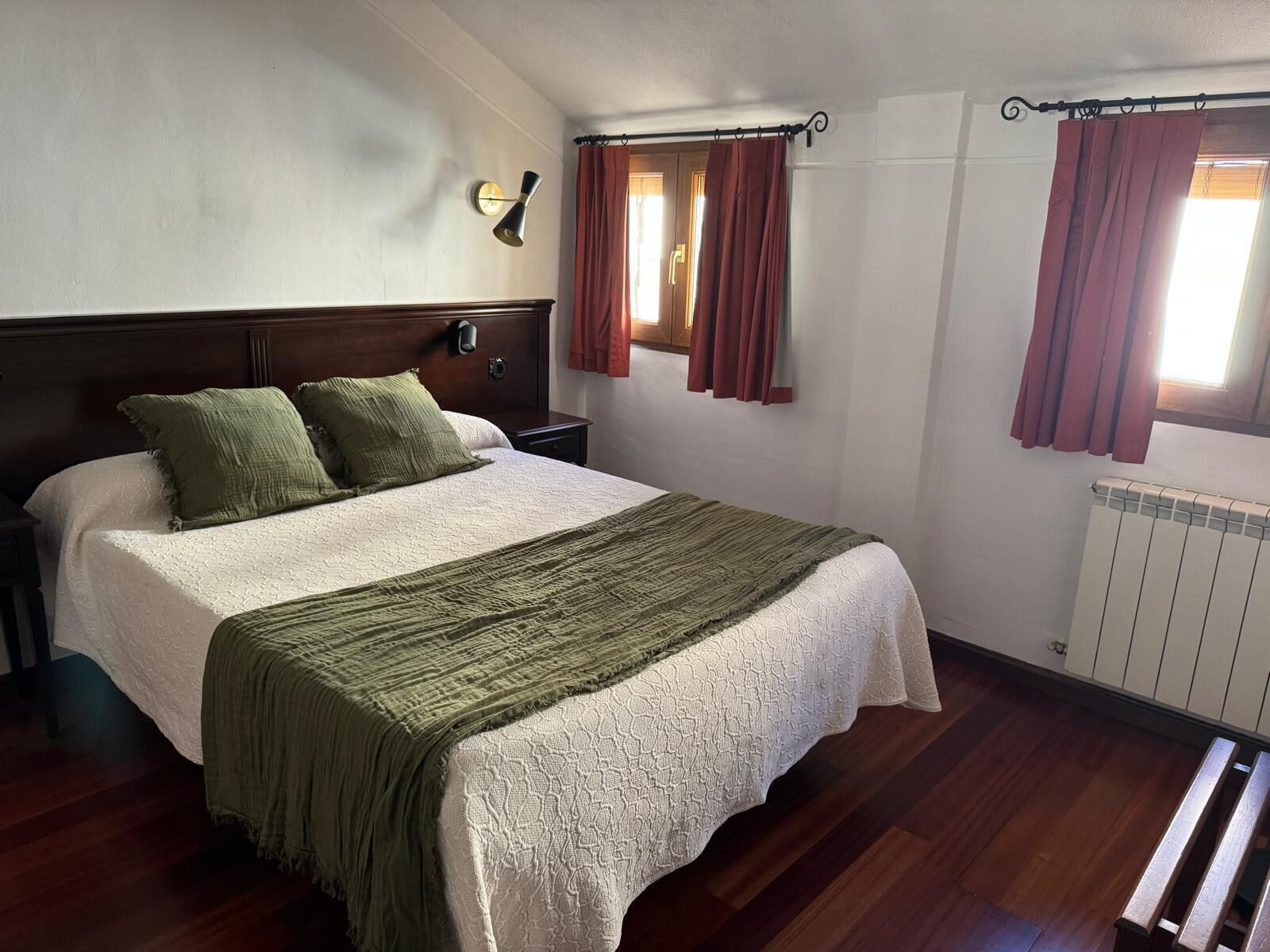 Un dormitorio con una cama grande, cabecera de madera, dos mesitas de noche, una lámpara de pared y una ventana con cortinas.