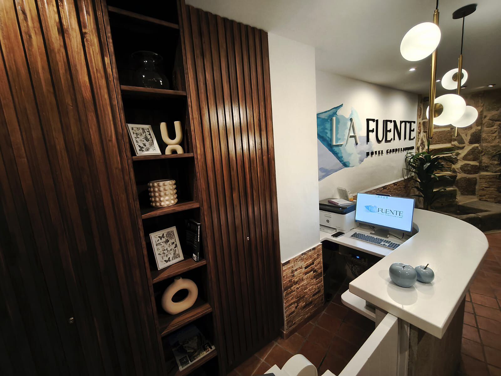 Una recepción con un ordenador portátil, una estantería de madera con objetos decorativos y una pared con el texto "LA FUENTE".
