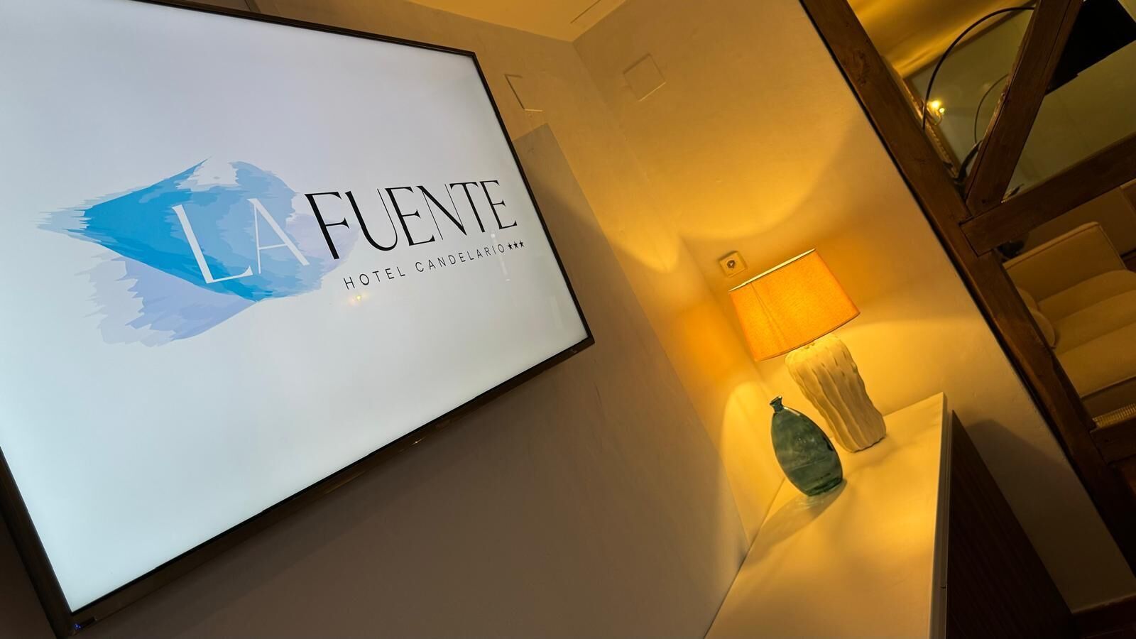 Un letrero en marco con el texto "LA FUENTE HOTEL CANDELARIO", una lámpara y un florero decorativo en una pared.