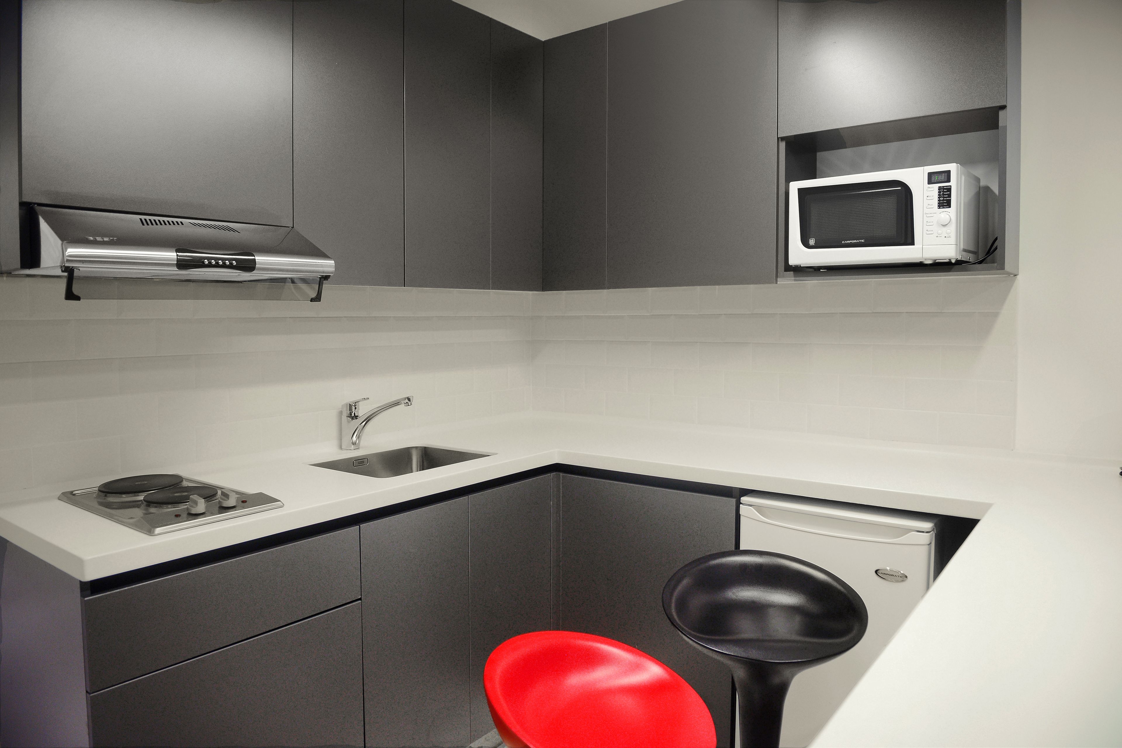 Une cuisine compacte équipée d’un four à micro-ondes encastré, d’une cuisinière à gaz et d’un tabouret de bar rouge.