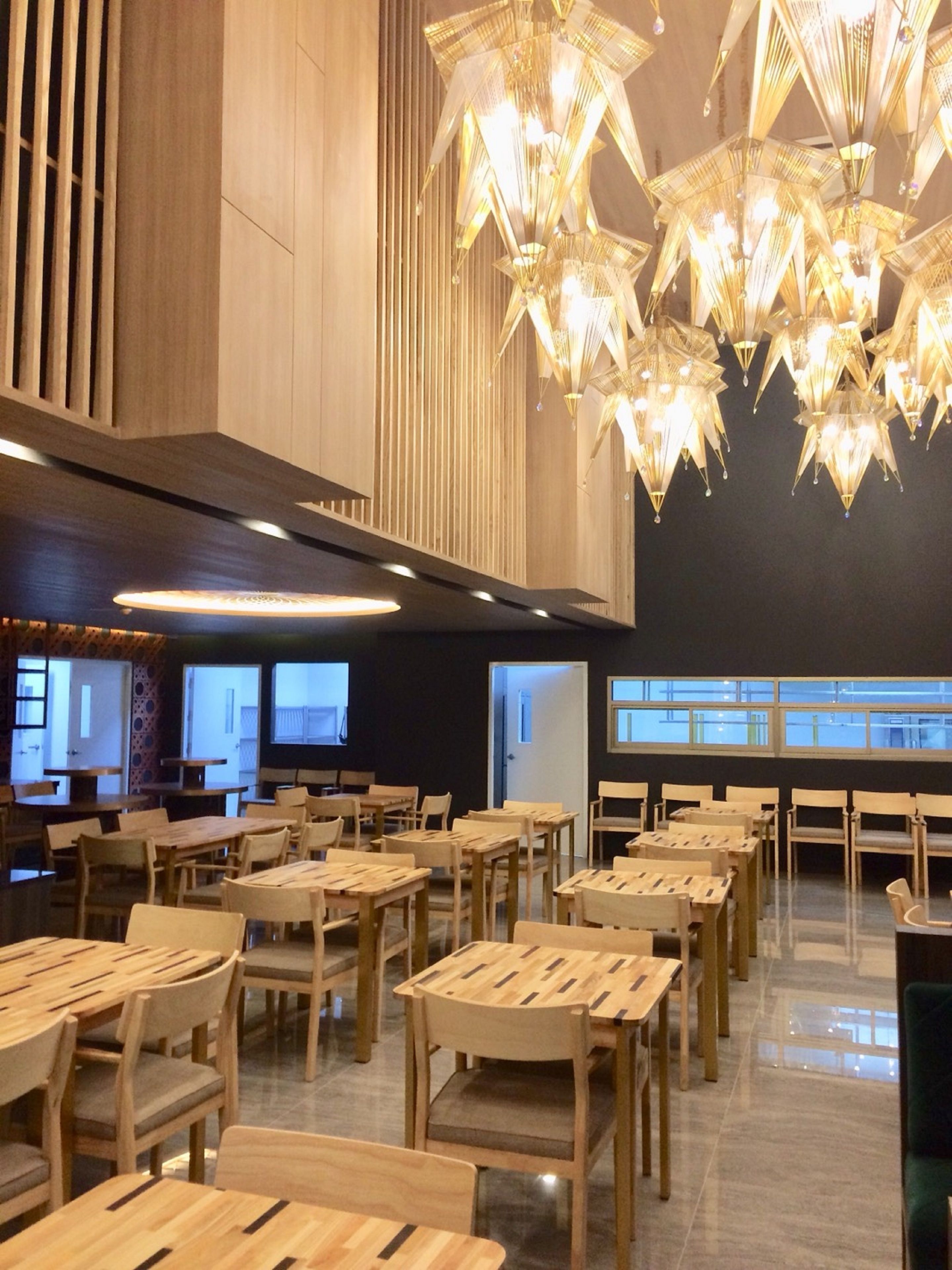 A spacious dining area with multiple wooden tables and chairs, a modern chandelier, and large windows allowing natural light.