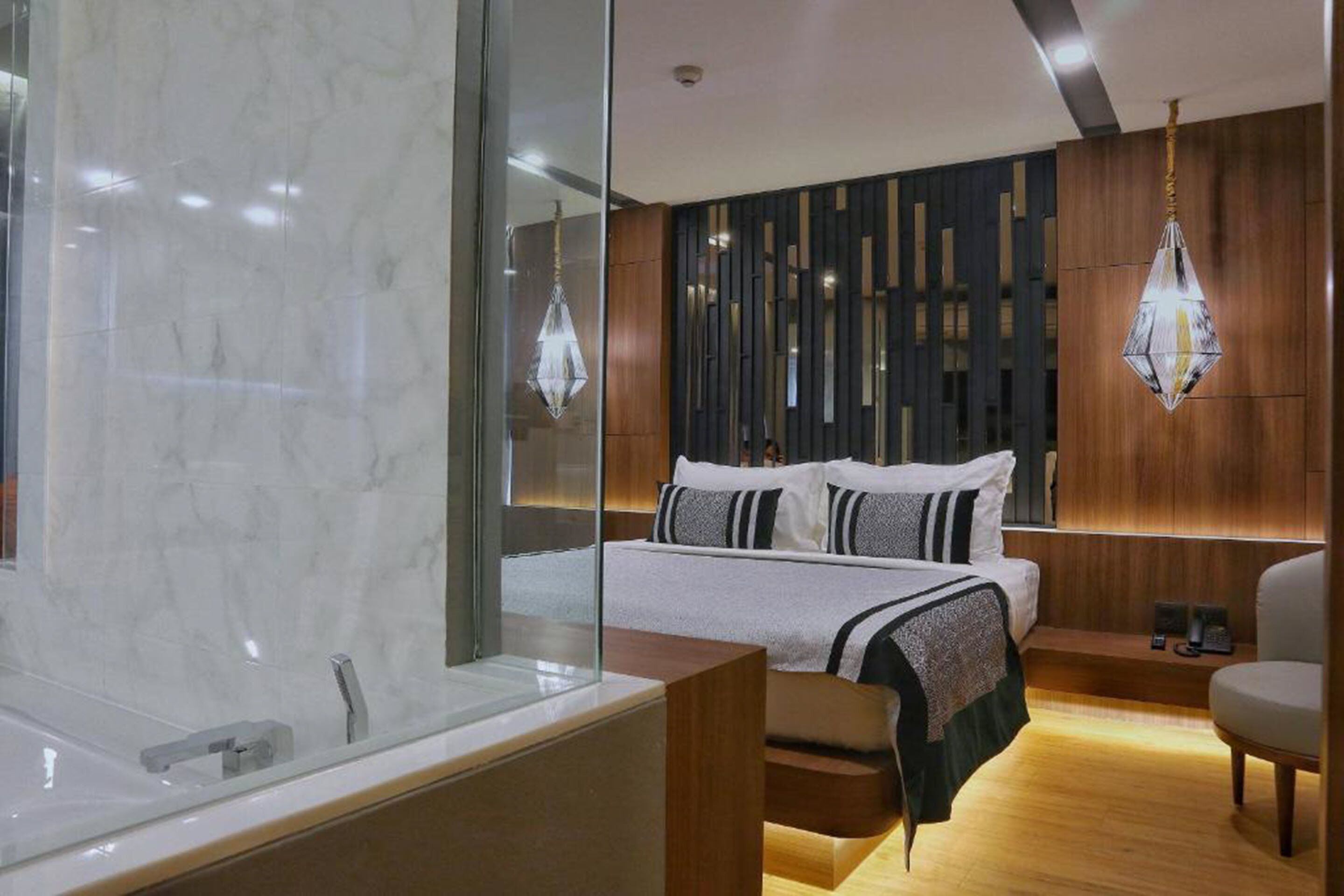 A modern hotel room with a large bed, a wooden side table, a chair, a glass shower enclosure, and a chandelier.
