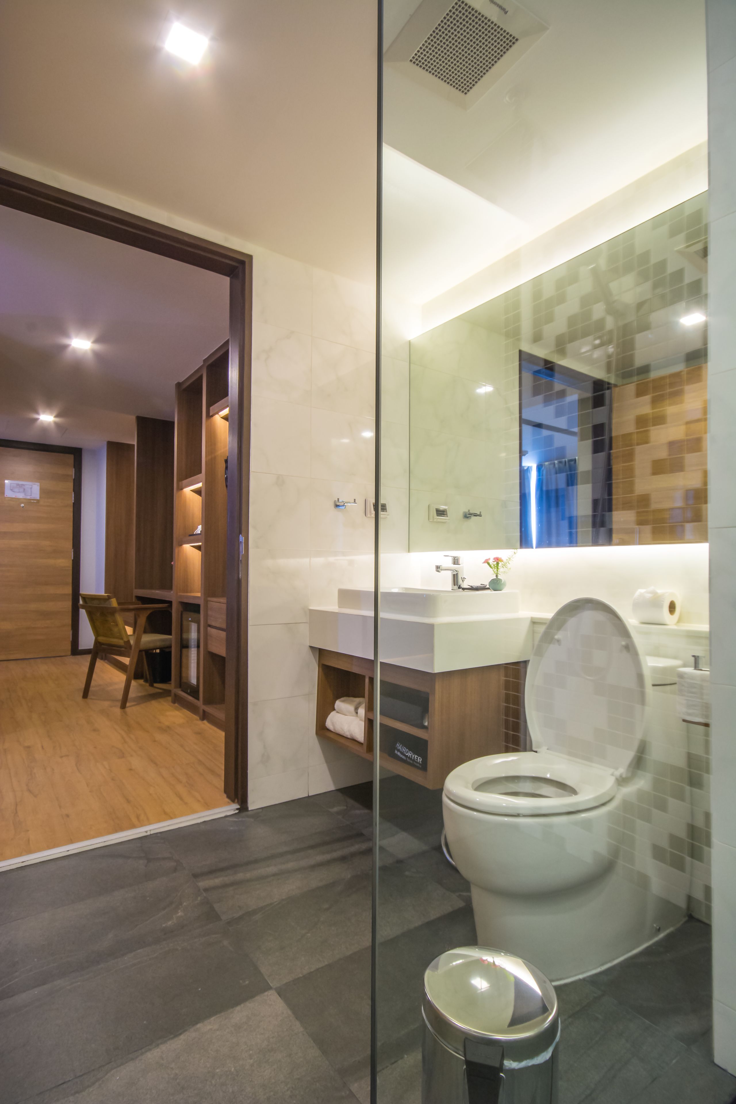 Un baño moderno con separador de vidrio, una silla de madera, un inodoro blanco y un tocador con espejo.