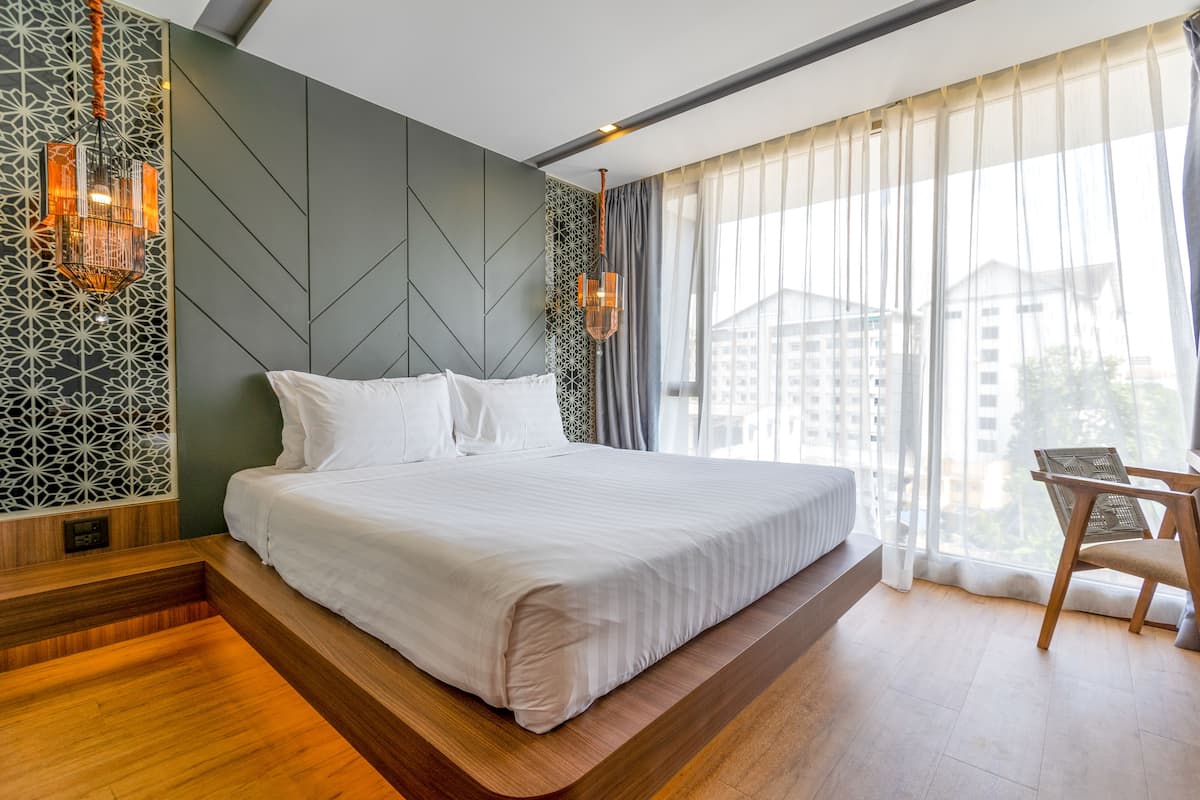 A modern hotel room with a large bed, a wooden platform, a chair, and a view of buildings through sheer curtains.