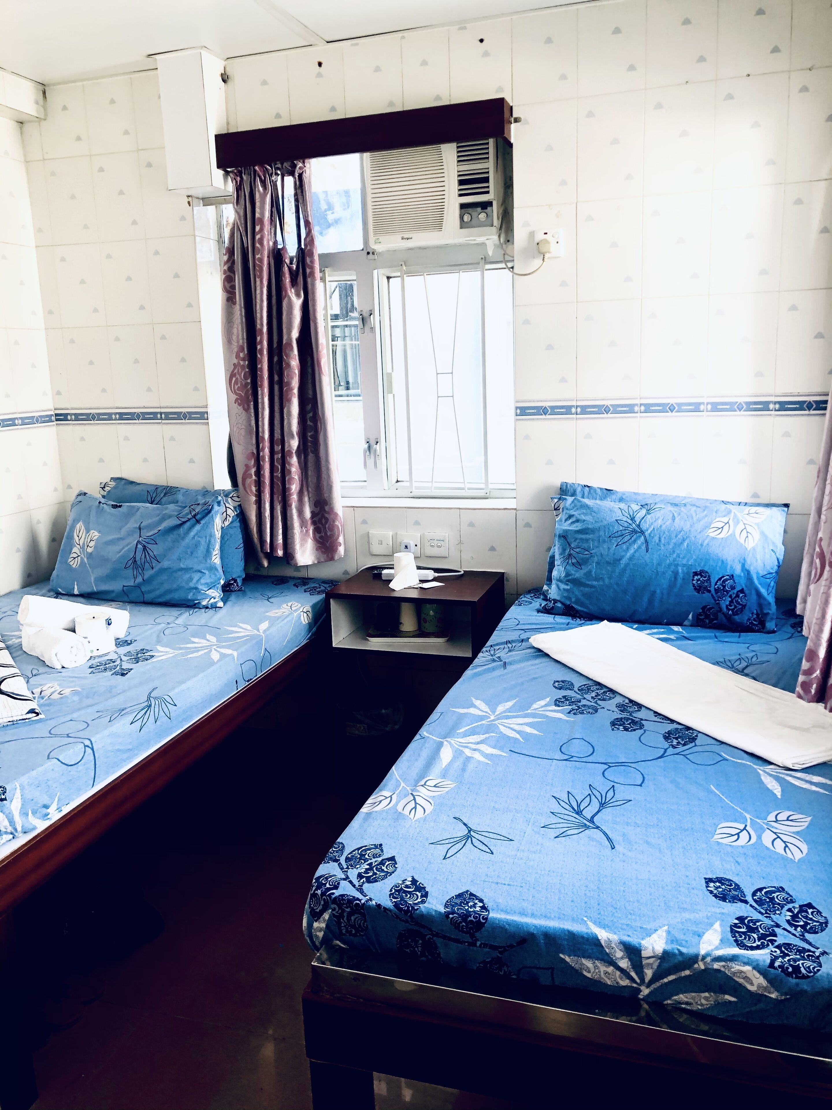 Ein Zimmer mit zwei Betten, beide mit blumigem, blauem Überwurf und passenden Kissen. Dazwischen steht ein Nachttisch mit Taschentuchbox und Lampe.