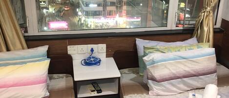 A hotel room with a bed, a small table with a remote control, and a view of a busy street at night.