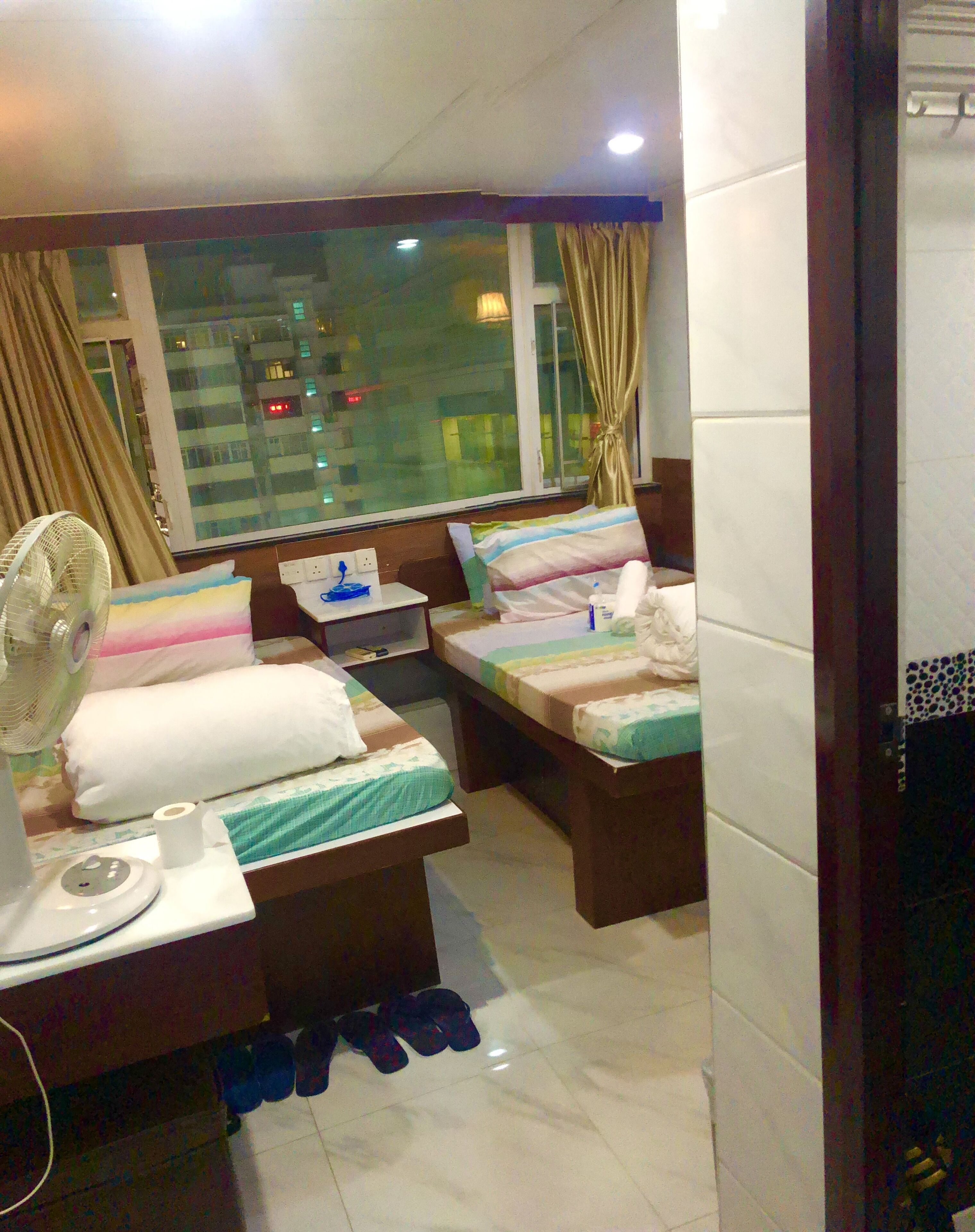A hotel room with two beds, a bedside table with a fan, a window with curtains, and a view of a cityscape at night.