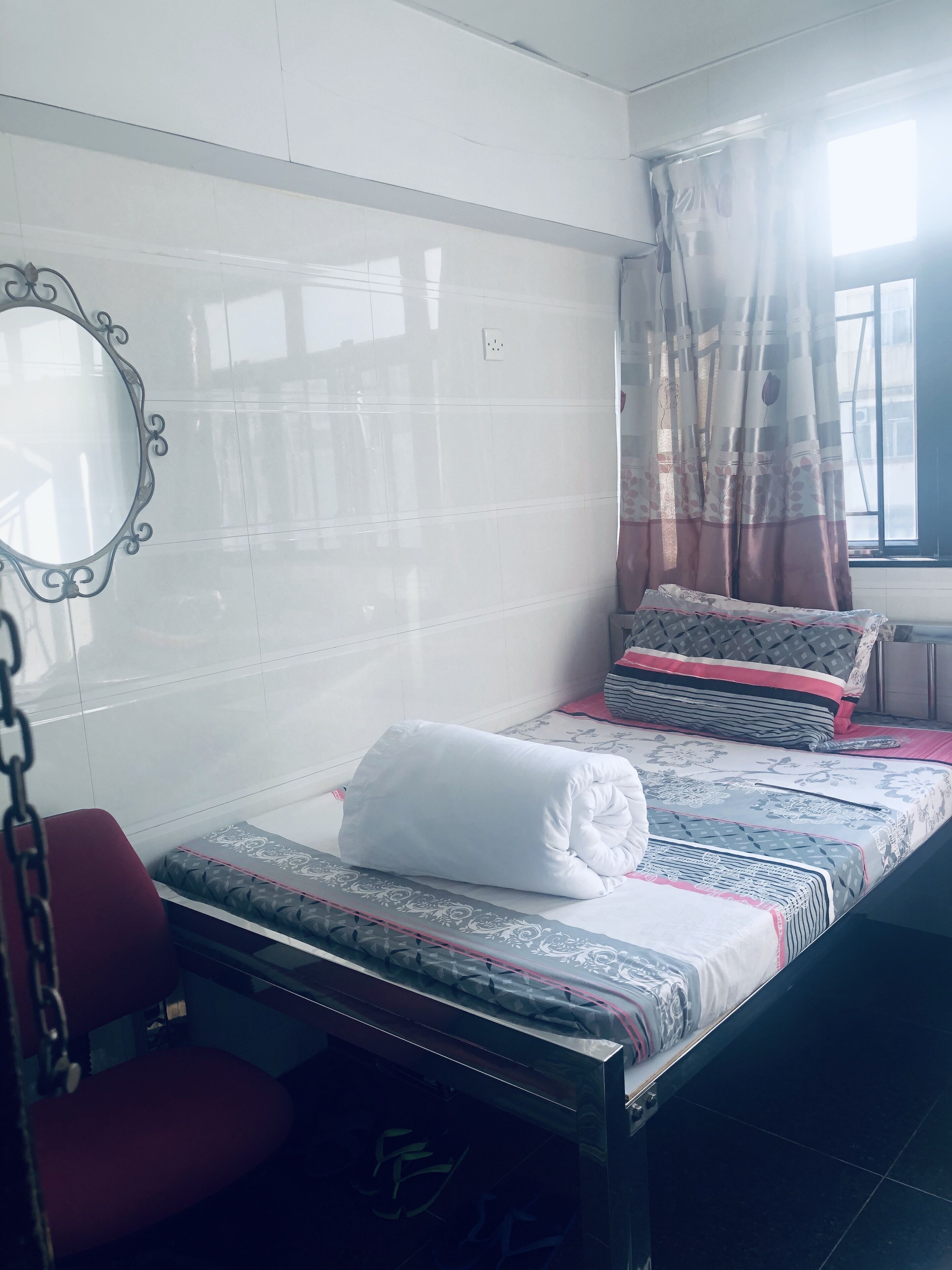 Un dormitorio pequeño y ordenado con cama, una silla, un espejo y una ventana con cortinas.