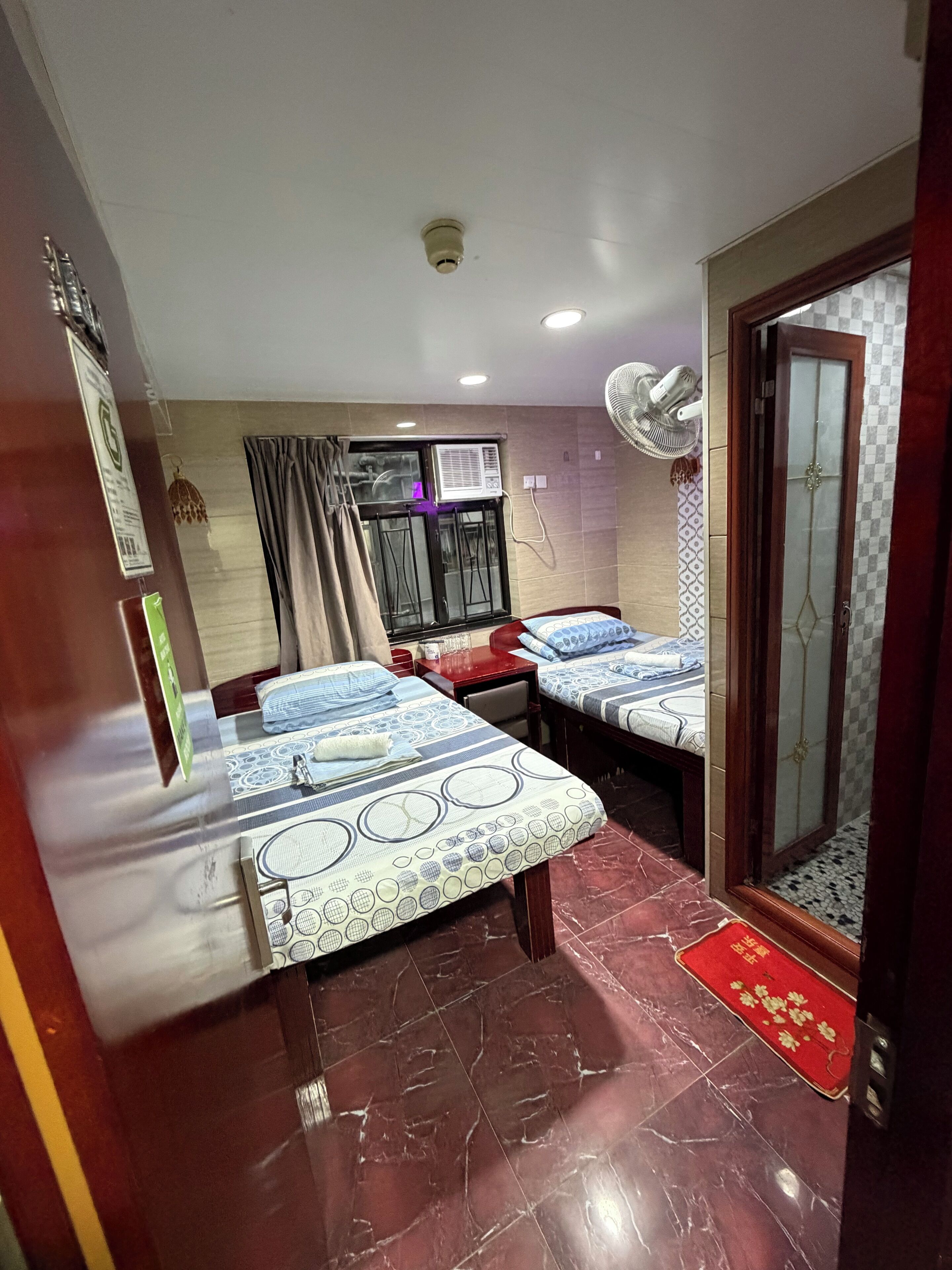 A hotel room with two beds, a window with curtains, an air conditioning unit, a ceiling fan, and a tiled floor.