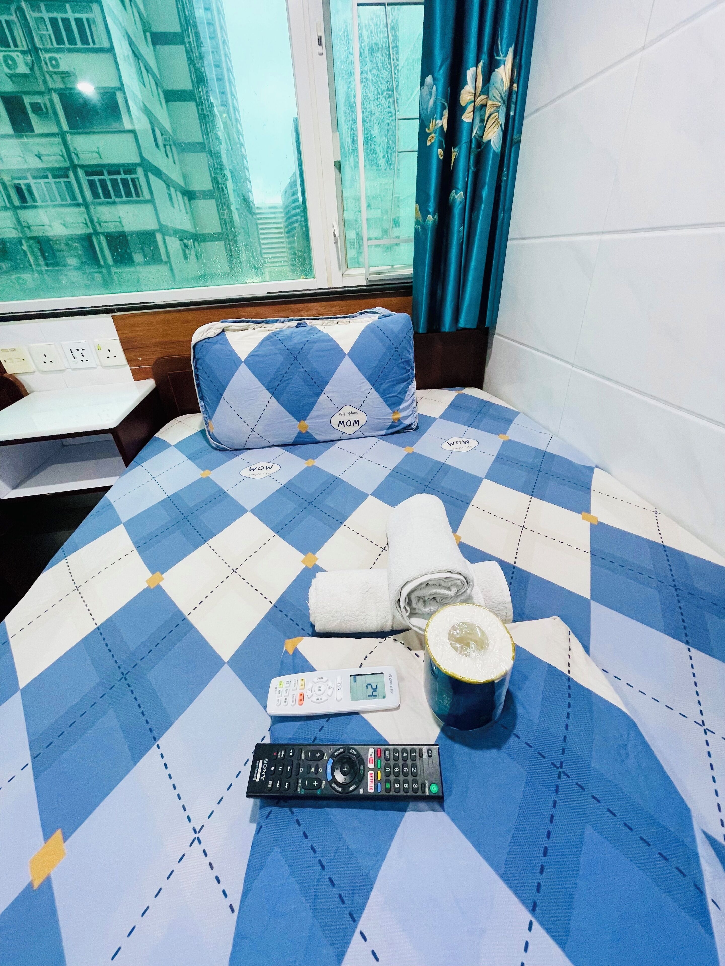 Un letto con copriletto a quadretti, un cuscino, un telecomando, un asciugamano arrotolato e una tazza.