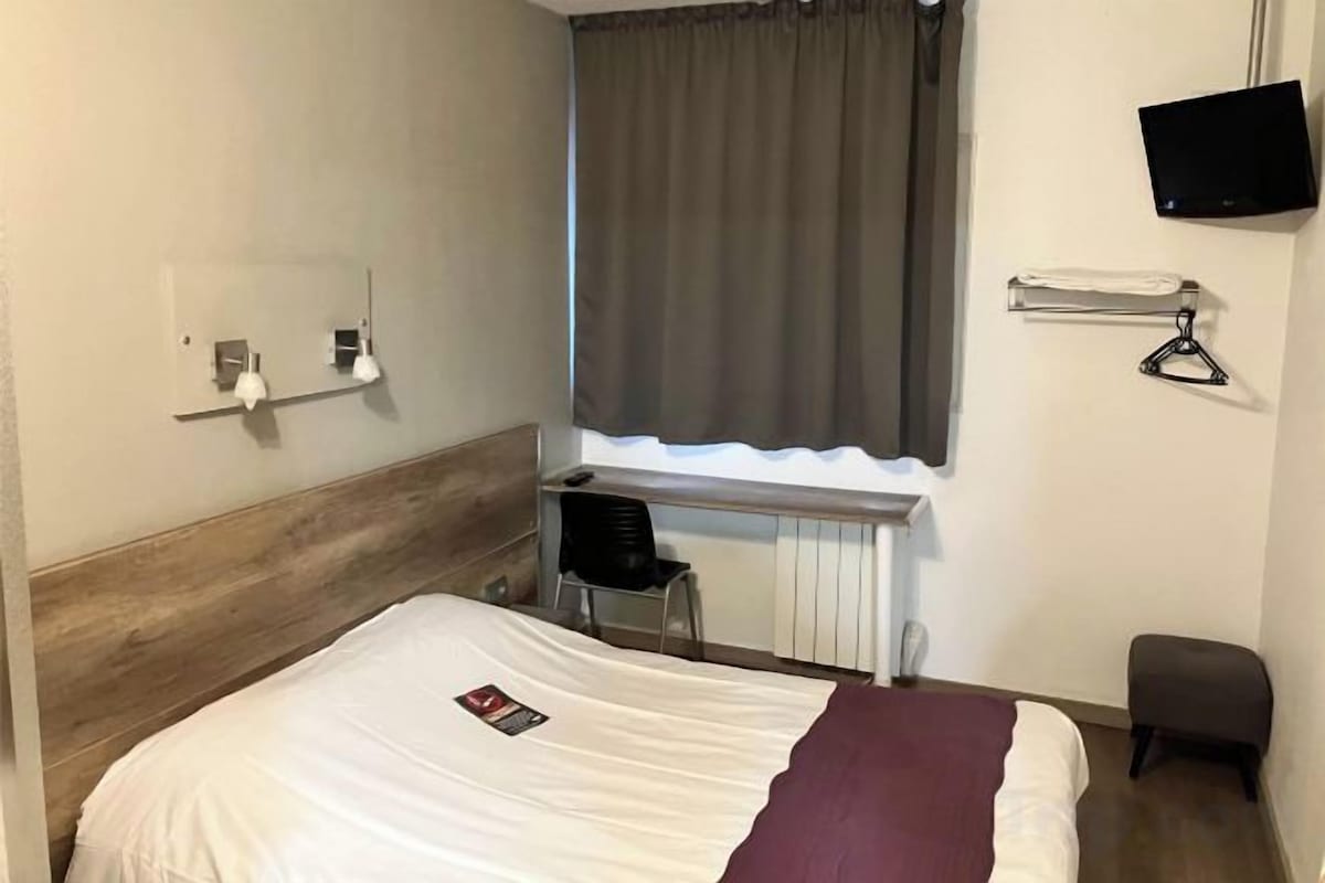 Une chambre d’hôtel avec un lit, un bureau, une chaise, une télévision et une fenêtre avec des rideaux.