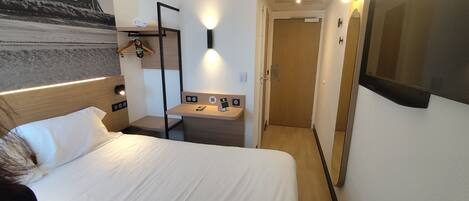 Een hotelkamer met een bed, een nachtkastje met oplaadpunt, een spiegel en een deur naar een gang.