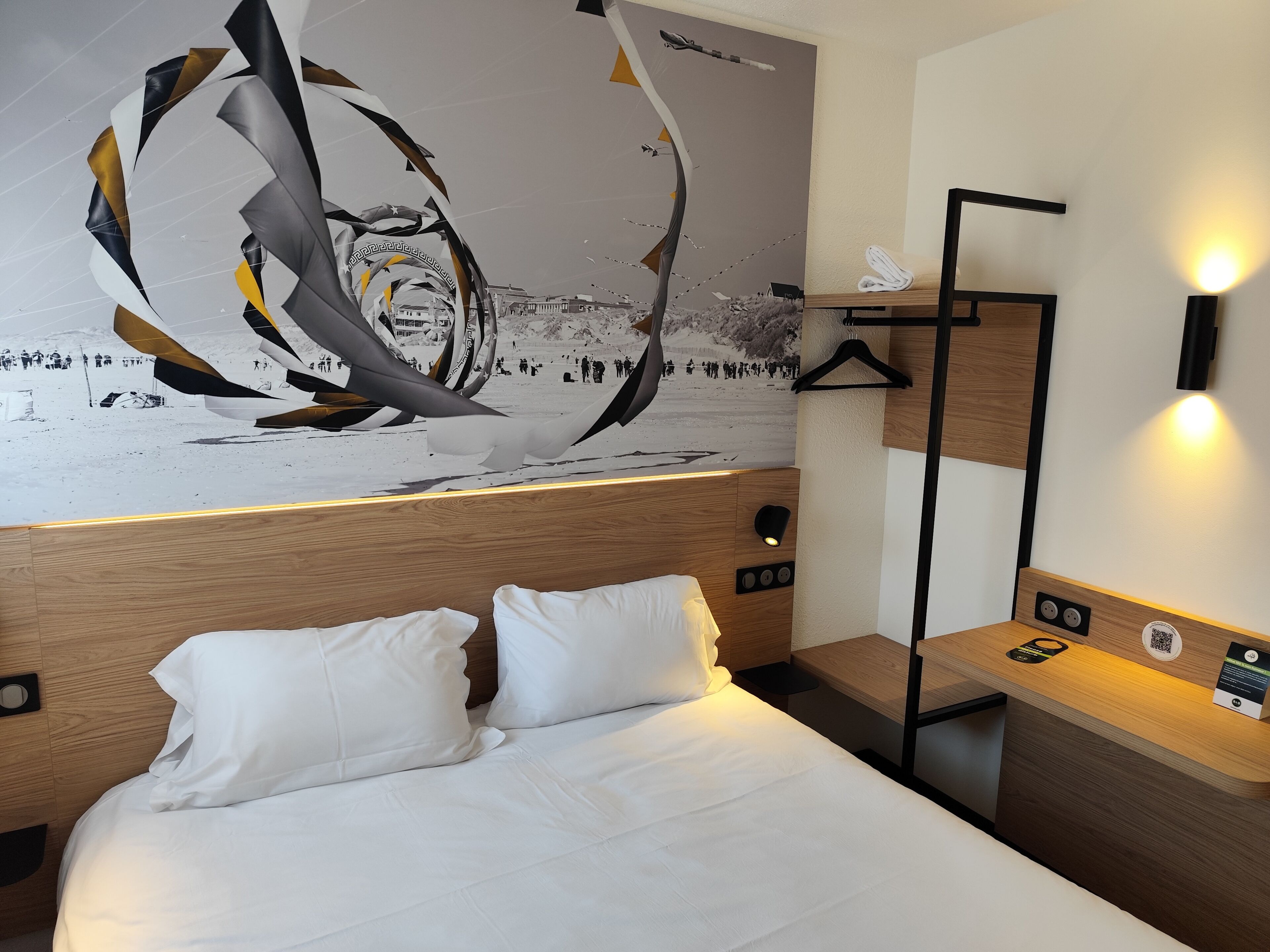Une chambre d’hôtel comprenant un lit, une table de chevet, une lampe fixée au mur et une œuvre d’art représentant un paysage enneigé.