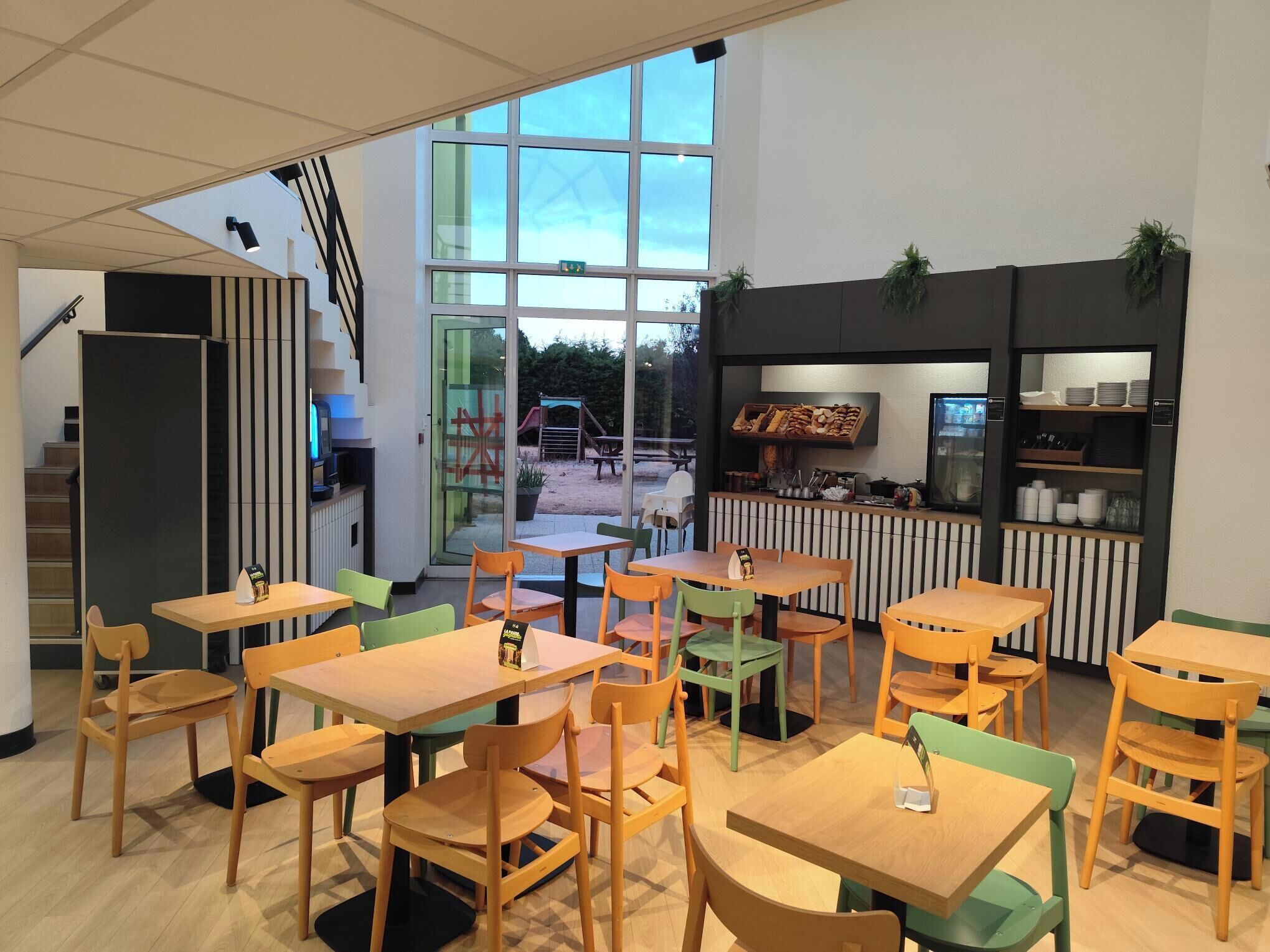 A modern indoor dining area with wooden tables and chairs, a kitchen counter with food items, and a view of a playground outside.