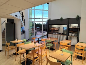 A modern indoor dining area with wooden tables and chairs, a kitchen counter with food items, and a view of a playground outside. - B&B HOTEL Berck-sur-Mer Rang-du-Fliers (Rang-du-Fliers)