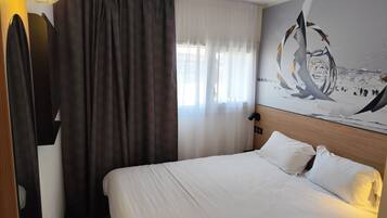 A hotel room with a bed, curtains, a wall-mounted lamp, and a large abstract painting.