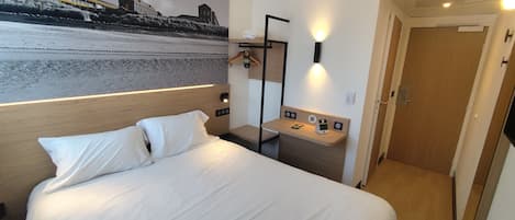 Una habitación de hotel con una cama, una mesita de noche, un espejo, una lámpara de pared y una puerta.