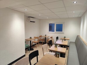 Een restaurantinterieur met meerdere tafels en stoelen, een raam met blauwe gordijnen en een airconditioner aan de muur.