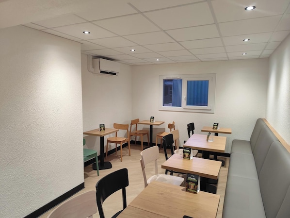 Interior de un restaurante con varias mesas y sillas, una ventana con cortinas azules y un equipo de aire acondicionado en la pared.