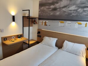 Hotelkamer met een bed, nachtkastje met lamp, wandplank en uitzicht op een strand met strandhutten.