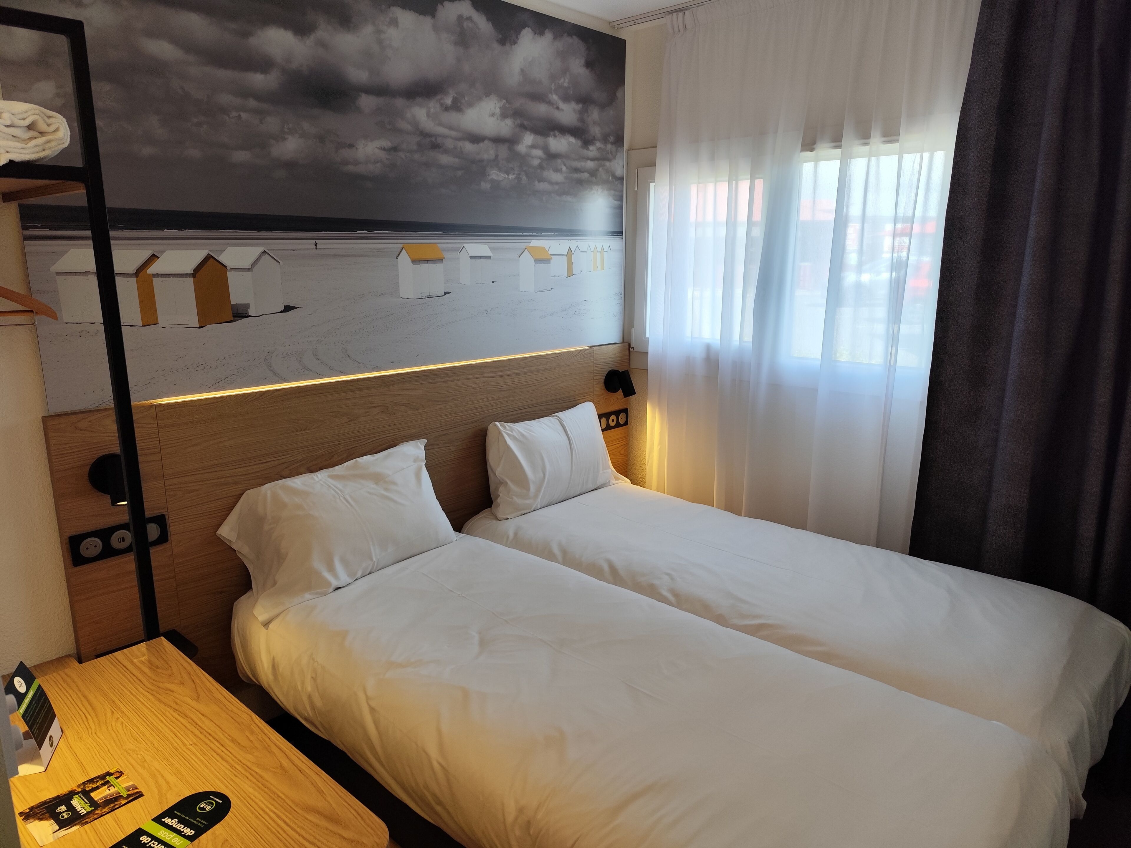A hotel room with a bed, a nightstand, a wall mural, and a window with curtains.