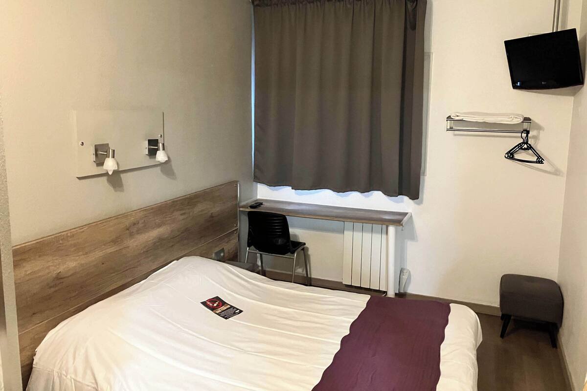 Une chambre d’hôtel avec un lit, un bureau, une chaise, une télévision et une fenêtre avec des rideaux.