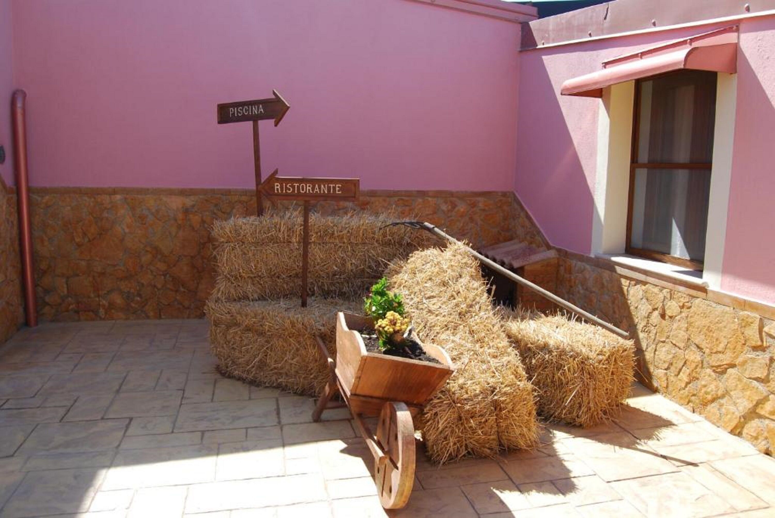 A rustic outdoor area with hay bales, a wooden wheelbarrow with a potted plant, and directional signs for 'Piscina' and 'Ristorante'.