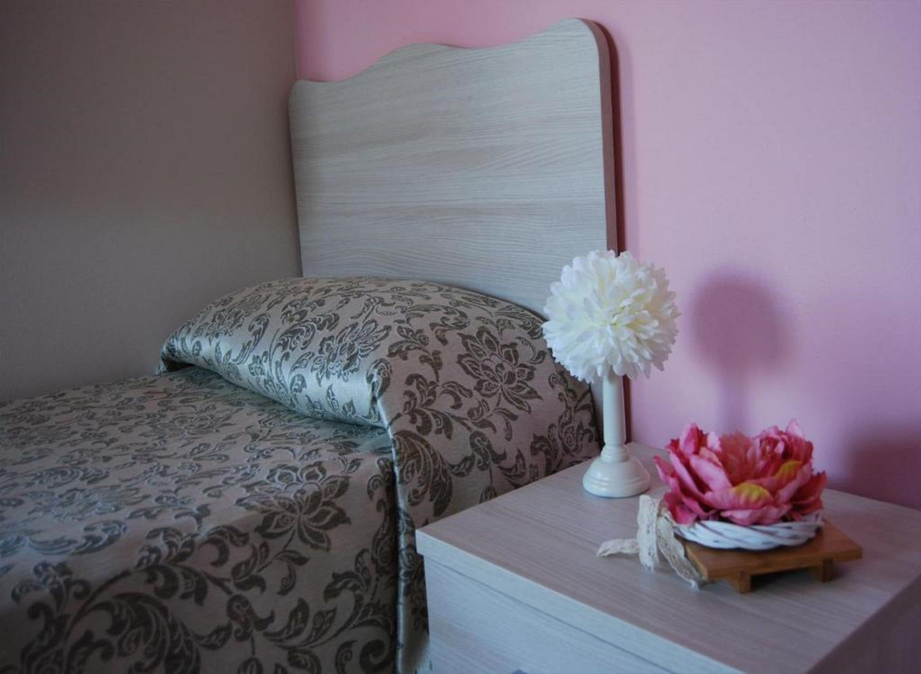 Un lit bien fait, avec une literie à motifs, une table de chevet ornée d’un vase de fleurs et un mur peint en rose.