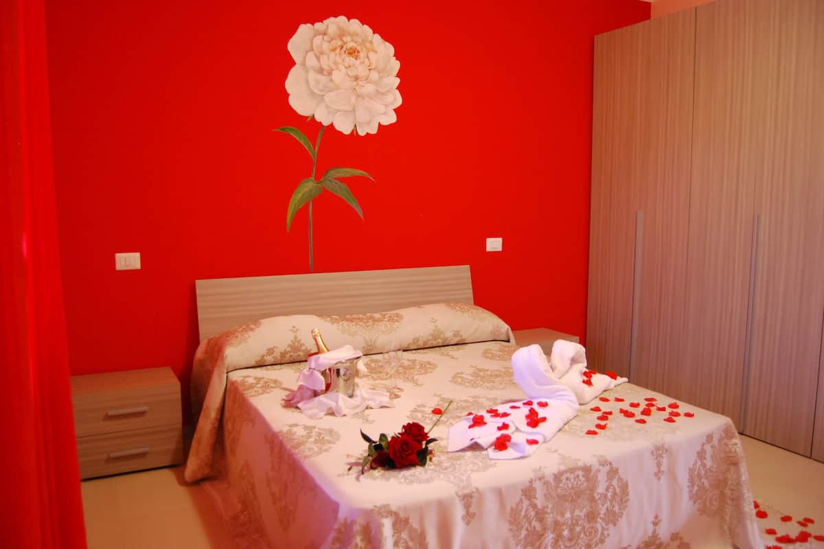 Une chambre avec un mur rouge, un grand tableau représentant une fleur blanche, un lit recouvert d’une courtepointe blanche et dorée, et des pétales de rose éparpillés sur le sol.