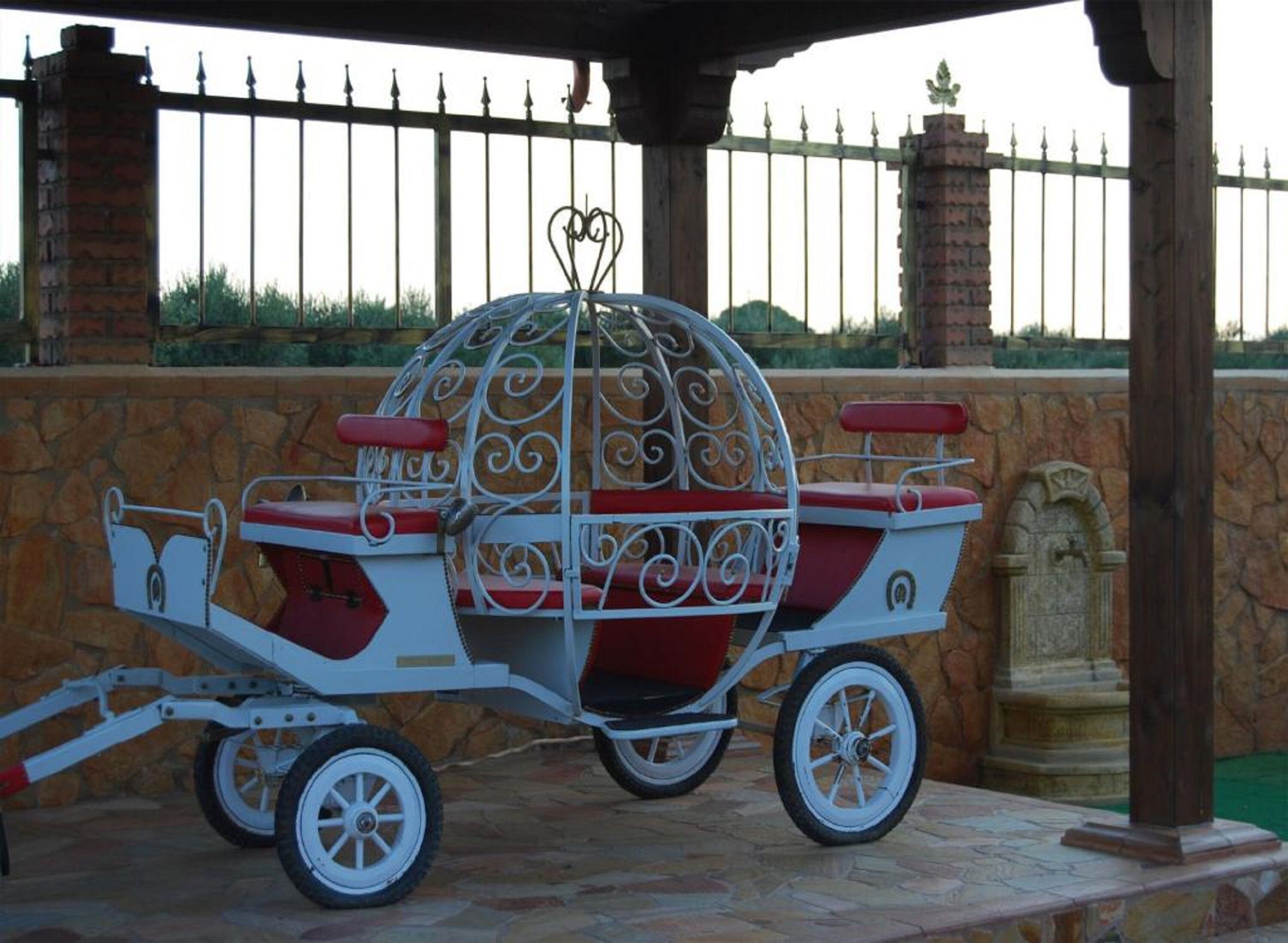 A carriage with ornate metalwork and red cushions, parked under a wooden structure with stone walls and a metal fence in the background.