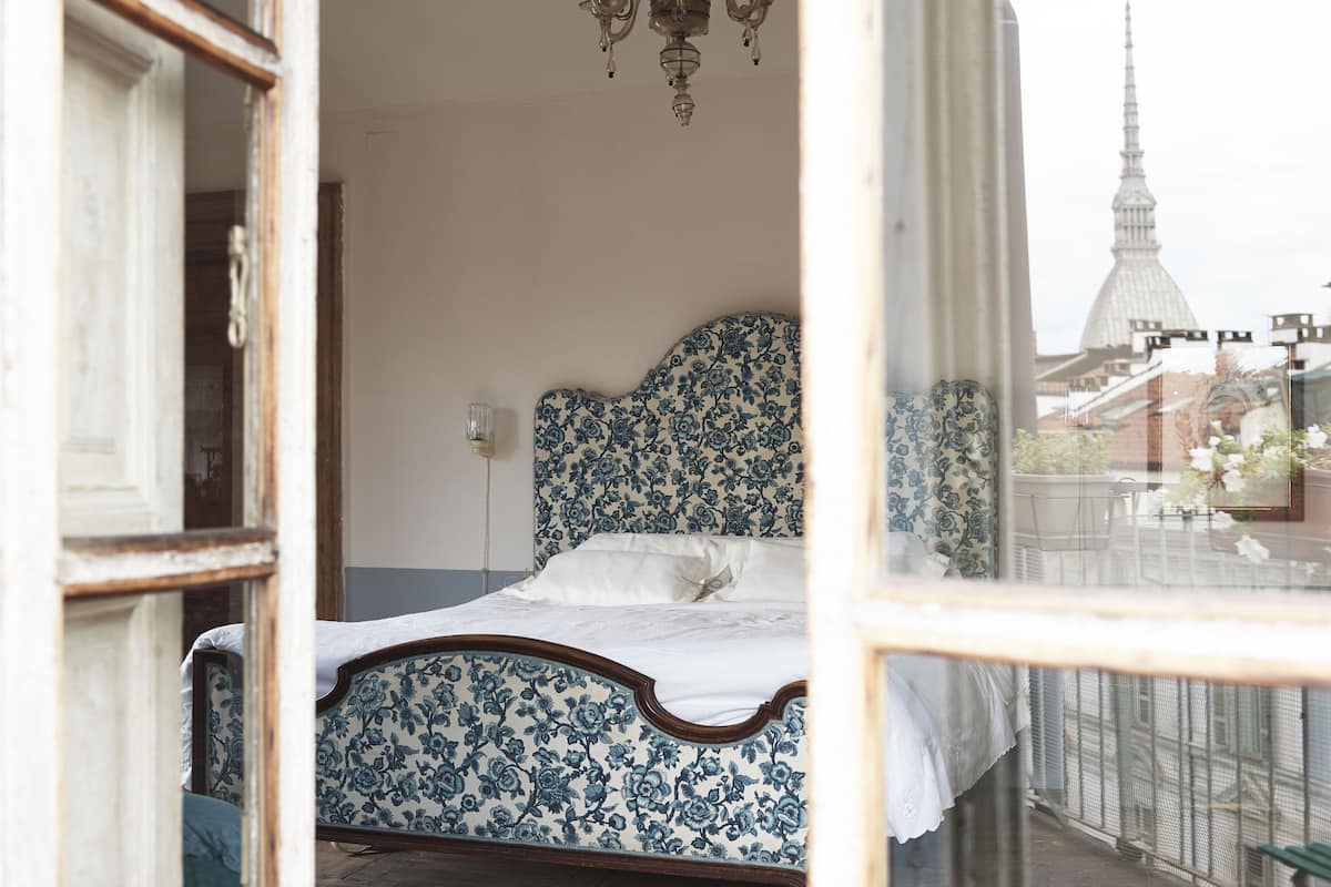 A bedroom with a floral-patterned bed, a chandelier, and a view of a cityscape through a window.