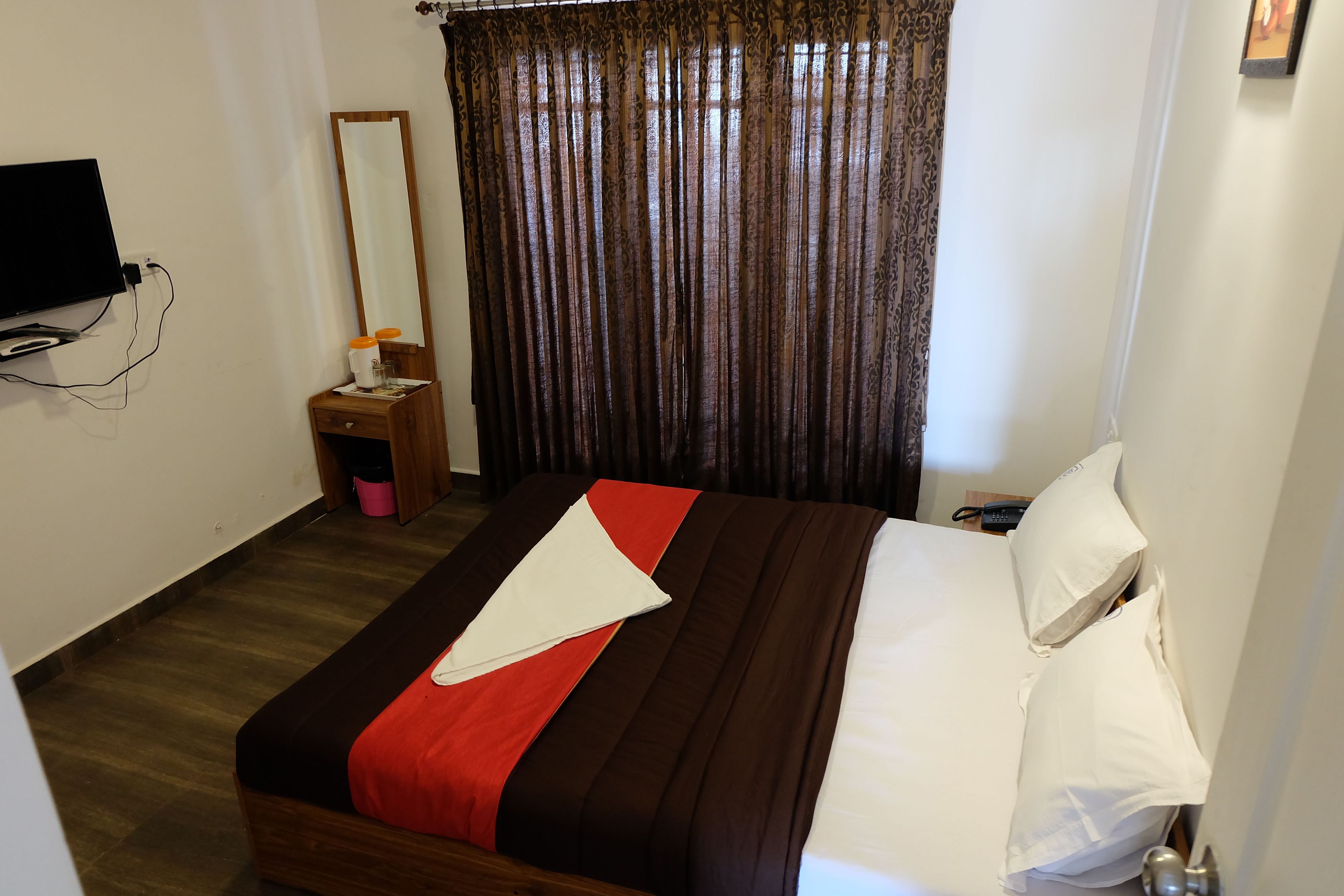 Un letto rifatto con una coperta rosso e marrone, un comodino in legno con una lampada e un grande specchio appeso al muro.
