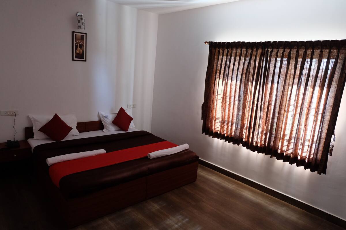 A bedroom with a large bed, a nightstand, a wall-mounted lamp, and a window with curtains.