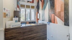 Ein Empfangstresen mit Holzoptik, ein Fenster mit Jalousien und eine Wand mit einem geometrischen Wandbild.