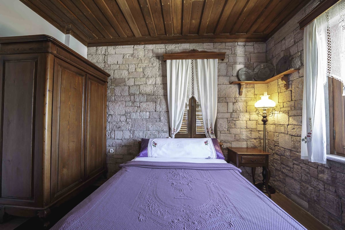 A bedroom with a stone wall, wooden ceiling, a bed with a purple bedspread, a nightstand with a lamp, and a wardrobe.