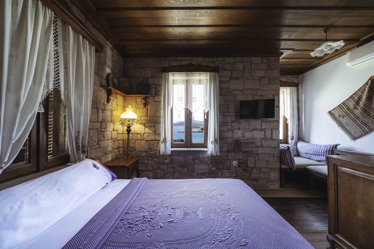 A bedroom with a stone wall, a bed with a purple blanket, a nightstand with a lamp, and a view of the sea through a window.