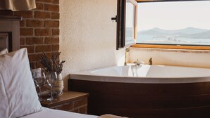 Un dormitorio con una bañera grande, una mesita de noche de madera, una almohada blanca y una ventana con vistas a las montañas.