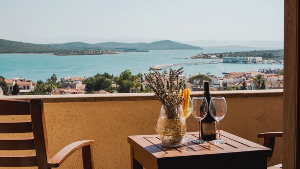Una mesa con una botella de vino y copas, un florero con flores y vistas a un pueblo costero y al mar.