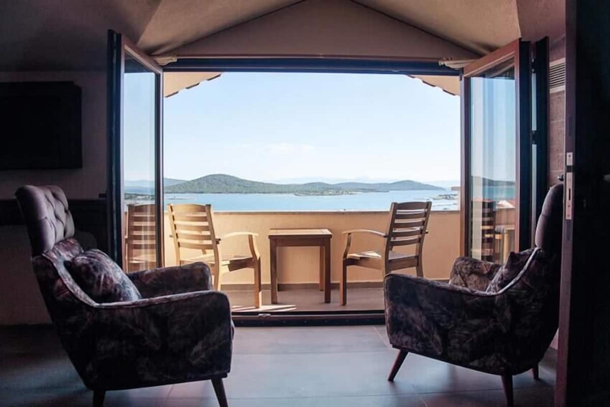 A balcony with two armchairs and a small table, overlooking a body of water and distant hills.