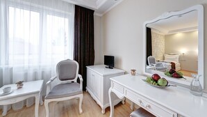 Quarto de hotel com toucador branco, uma cadeira, uma escrivaninha, uma televisão e um espelho grande.