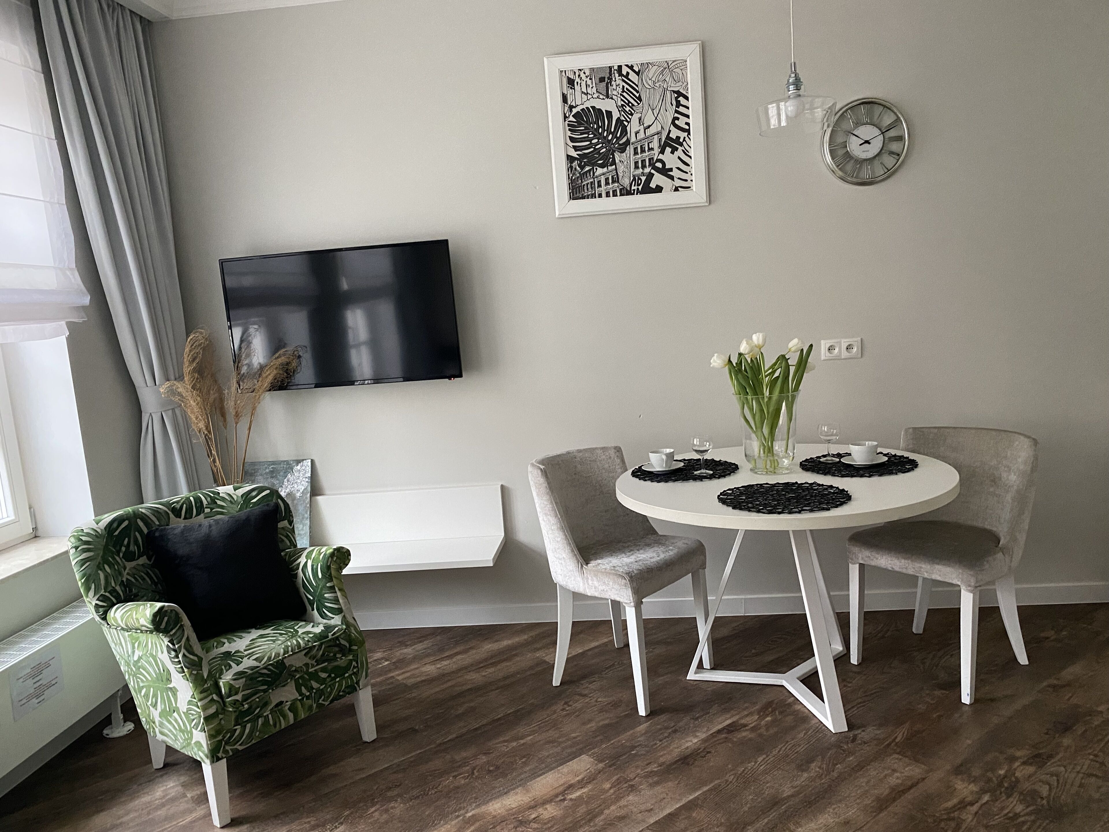 A modern living room with a flat-screen TV, a round dining table with four chairs, a green patterned armchair, and a wall-mounted clock.