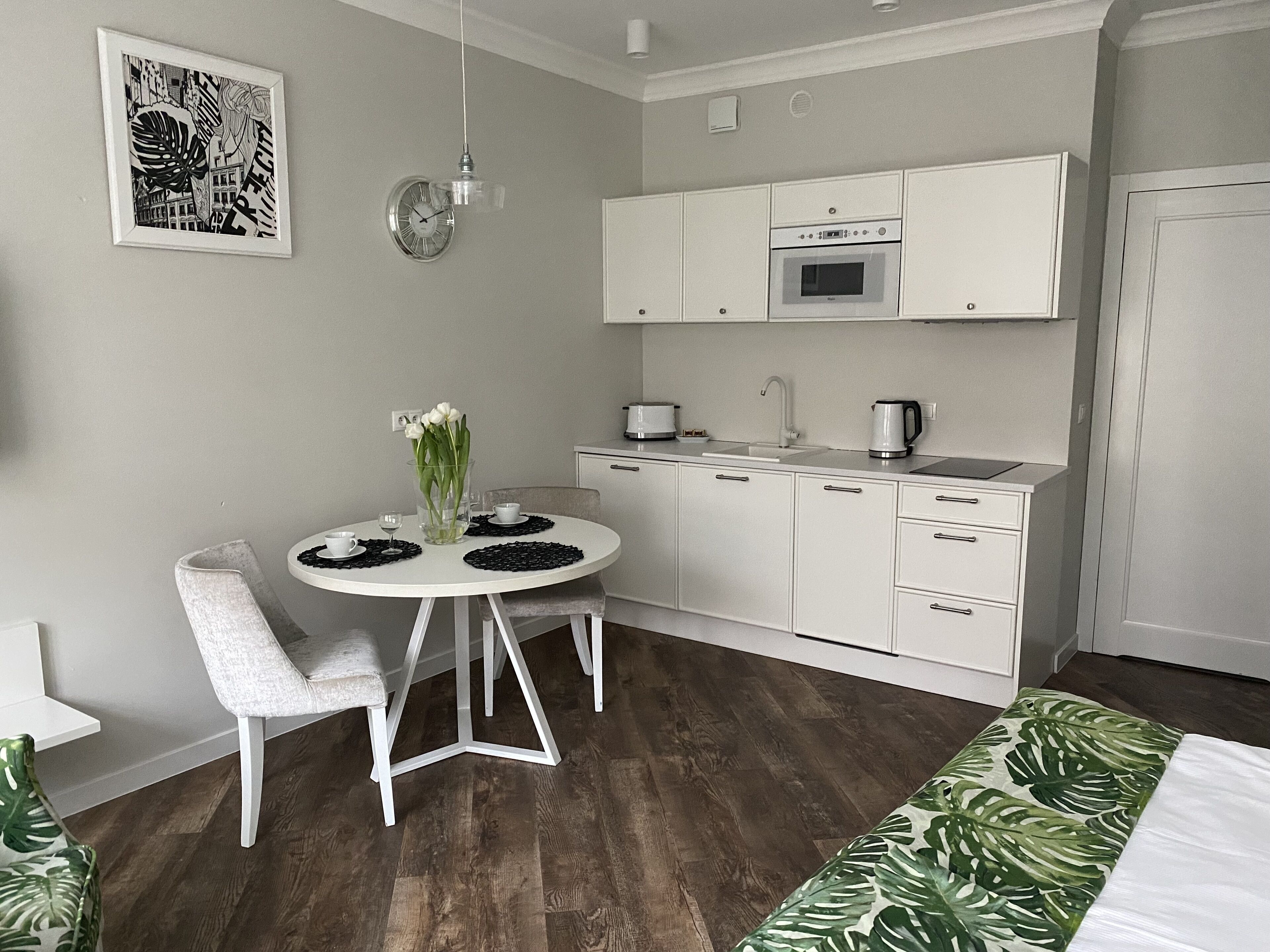 Una cocina compacta con gabinetes blancos, horno integrado y un lavamanos. Hay una mesa de comedor con sillas y un florero con flores.