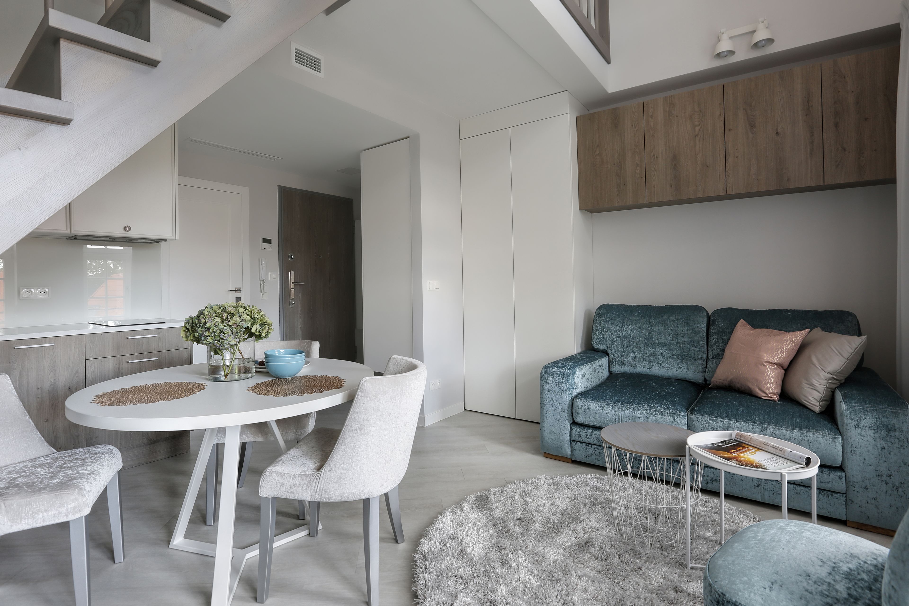 A modern living room with a round dining table, grey upholstered chairs, a blue sofa with cushions, and a fluffy grey rug.