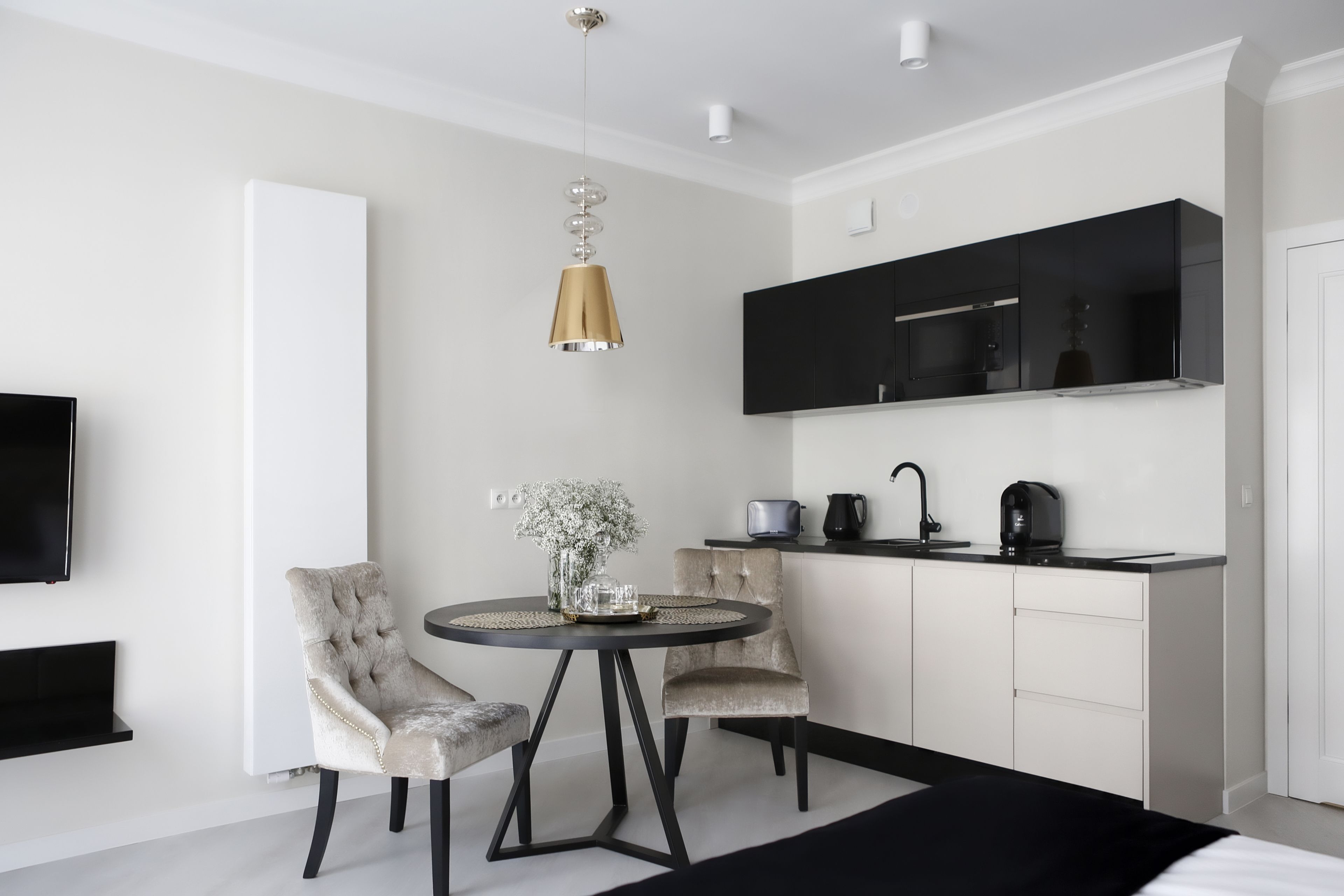 A modern, minimalist room with a round dining table, two upholstered chairs, a pendant light, a flat-screen TV, a black cabinet, and a white door.
