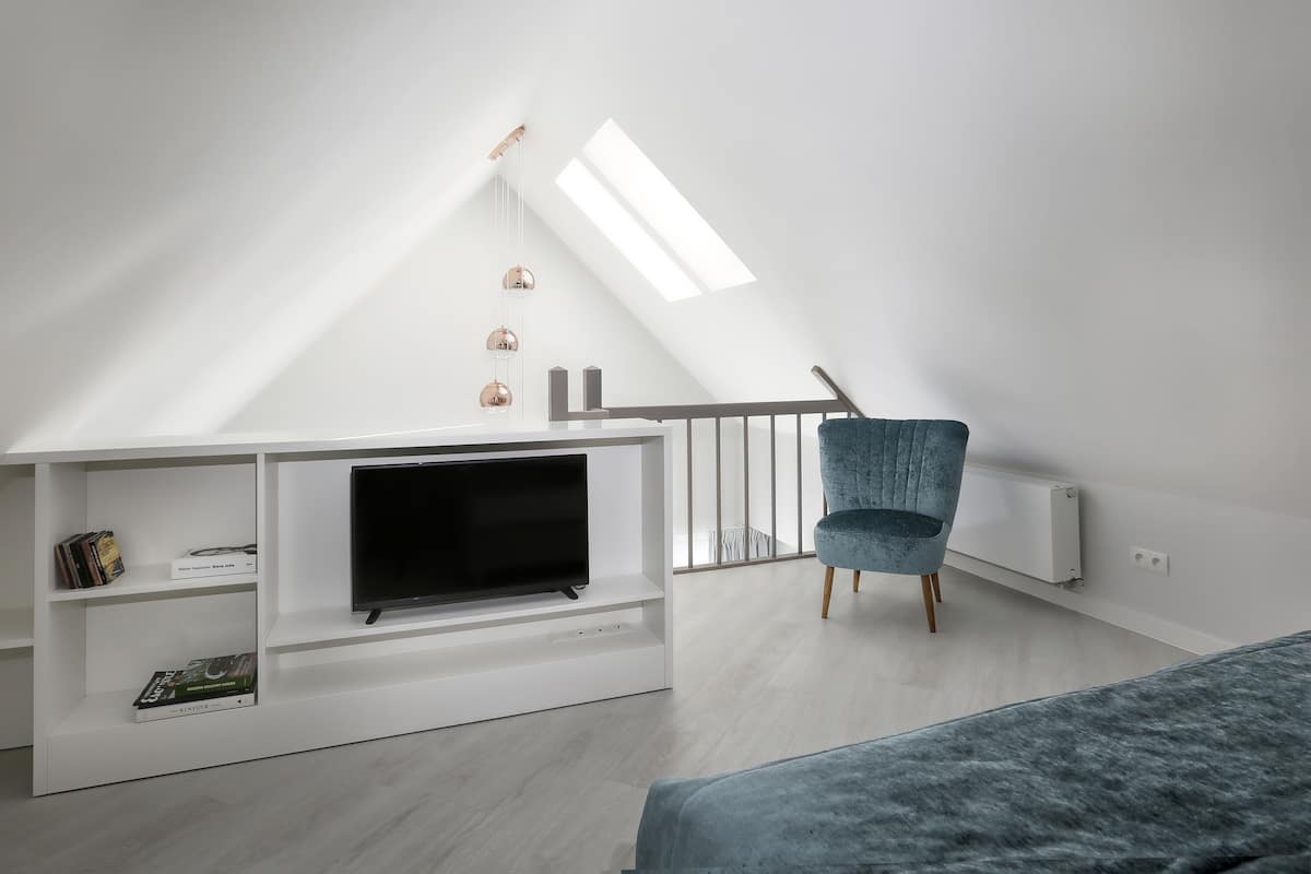 A modern loft bedroom with a sloped ceiling, a built-in TV, a blue chair, and a skylight.