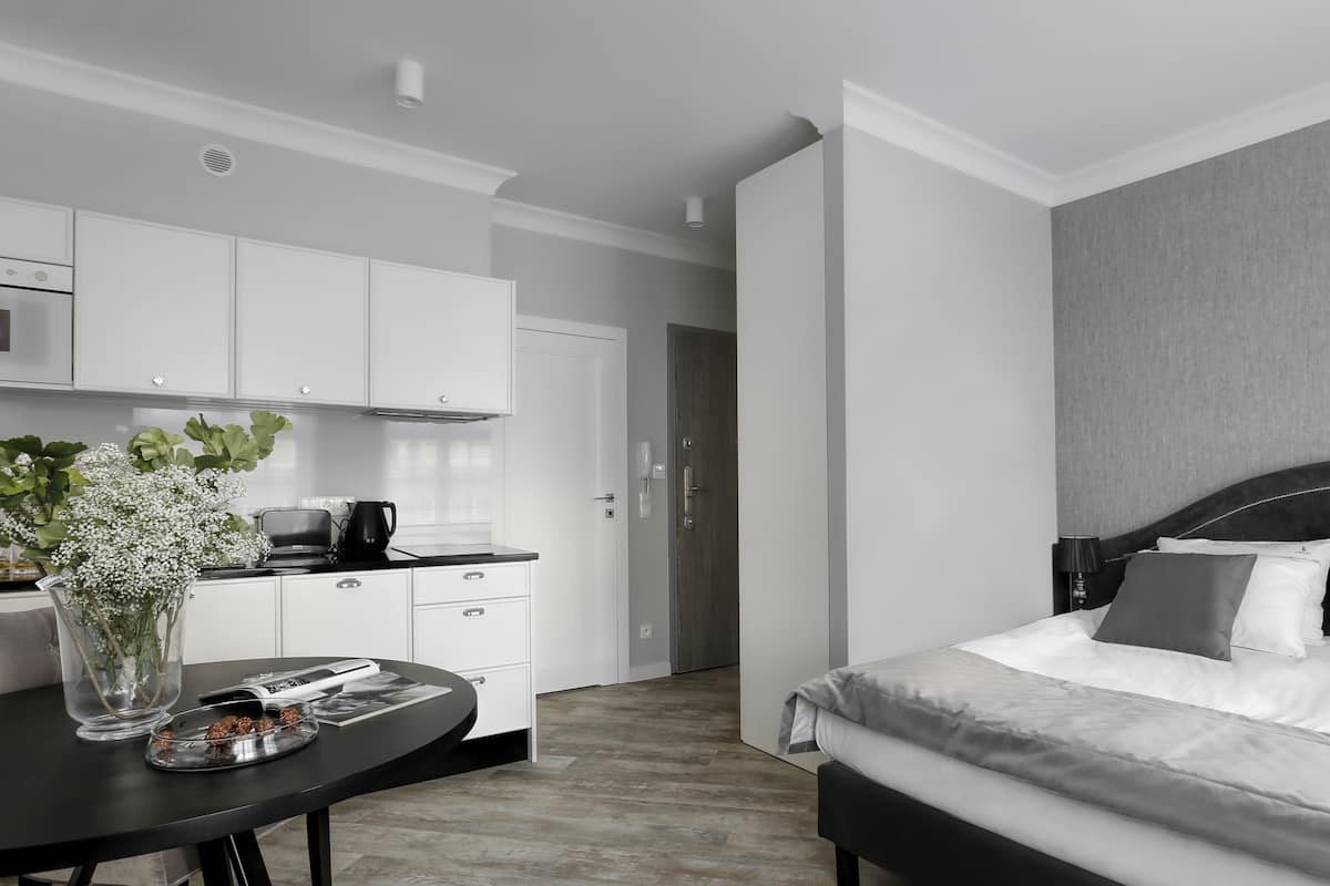 A modern hotel room with a bed, bedside table, and a kitchenette area.