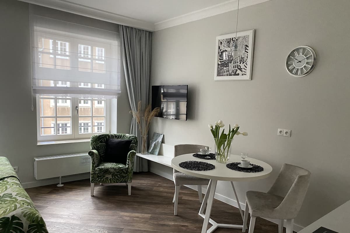 A modern living room with a dining table, chairs, a green patterned armchair, a large window with curtains, a flat-screen TV mounted on the wall, and a clock.